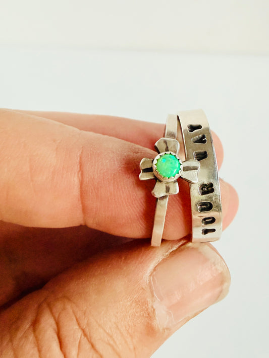 Two rings, one with a green gemstone and the other with engraved text, held between fingers against a white background.